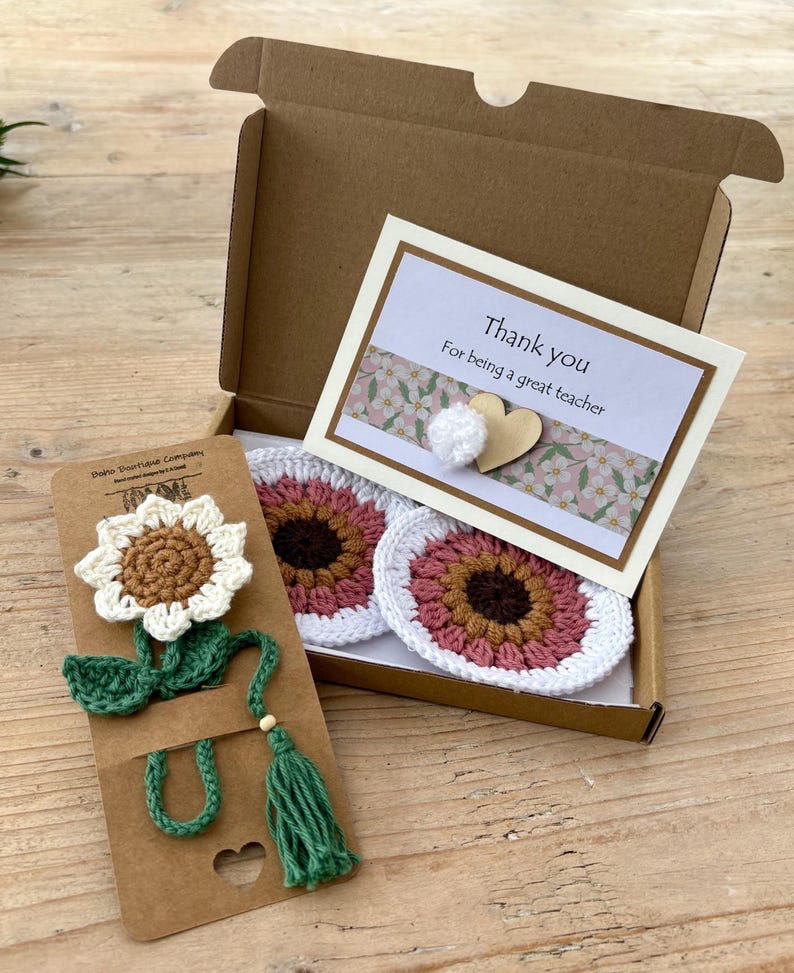 May include: A brown cardboard gift box containing two round crocheted coasters with pink and brown centres and white edges, and a card that reads Thank you For being a great teacher. A crocheted sunflower bookmark is attached to a brown card.