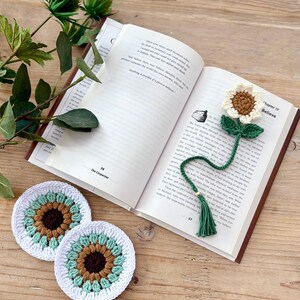 May include: A crochet sunflower bookmark with a green stem and tassel, placed inside an open book, alongside two round crochet coasters featuring a brown centre, light green petals, and a white border, all crafted with yarn in a floral design.