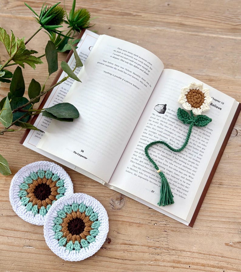 May include: A crochet sunflower bookmark with a green stem and tassel, placed inside an open book, alongside two round crochet coasters featuring a brown centre, light green petals, and a white border, all crafted with yarn in a floral design.