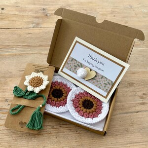 May include: A brown cardboard gift box containing two round crochet coasters with pink, brown, and white petals, and a crochet sunflower bookmark on a card. A white card with the text Thank you For helping me grow is attached with a heart.