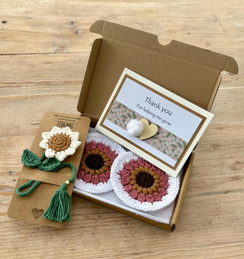 May include: A brown cardboard gift box containing two round crochet coasters with pink, brown, and white petals, and a crochet sunflower bookmark on a card. A white card with the text Thank you For helping me grow is attached with a heart.