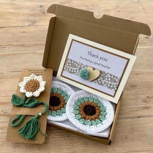 May include: A brown cardboard gift box containing two round crochet sunflower coasters and a card that reads Thank you For being a great teacher. Beside the box is a crochet sunflower bookmark with a green stem and tassel on a brown card.