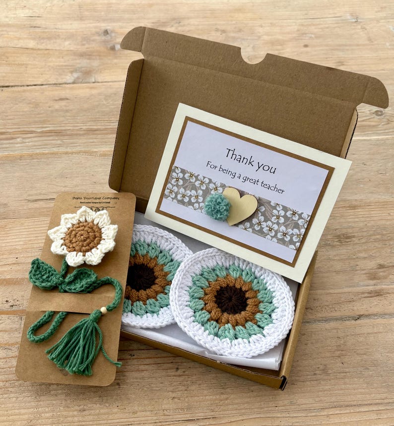 May include: A brown cardboard gift box containing two round crochet sunflower coasters and a card that reads Thank you For being a great teacher. Beside the box is a crochet sunflower bookmark with a green stem and tassel on a brown card.
