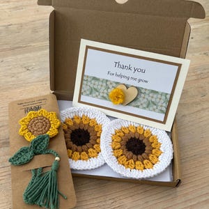 May include: A brown cardboard gift box containing two round crochet sunflower coasters with white borders, a crochet sunflower bookmark with a green tassel, and a white card with the text Thank you For helping me grow and a small wooden heart.