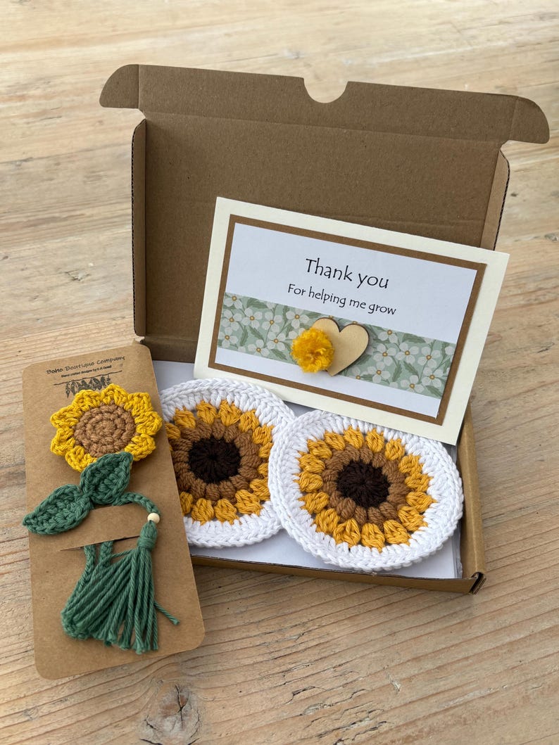 May include: A brown cardboard gift box containing two round crochet sunflower coasters with white borders, a crochet sunflower bookmark with a green tassel, and a white card with the text Thank you For helping me grow and a small wooden heart.