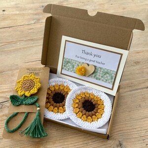 May include: A brown cardboard gift box containing two round white crochet coasters with yellow and brown sunflower patterns, and a matching crochet sunflower bookmark with a green stem and tassel on a brown card, plus a thank you note.