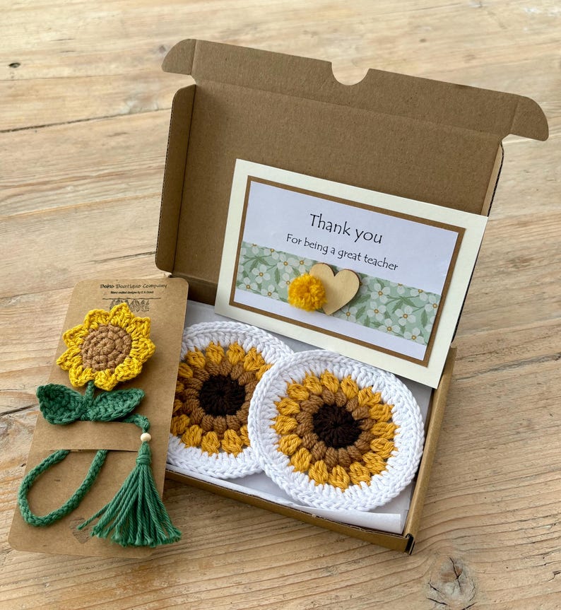 May include: A brown cardboard gift box containing two round white crochet coasters with yellow and brown sunflower patterns, and a matching crochet sunflower bookmark with a green stem and tassel on a brown card, plus a thank you note.