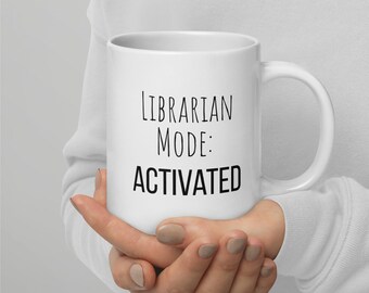 Puede incluir: Taza de cerámica blanca con el texto "Librarian Mode: ACTIVATED" en letras negras. La taza tiene un asa curva y la sostiene una persona.