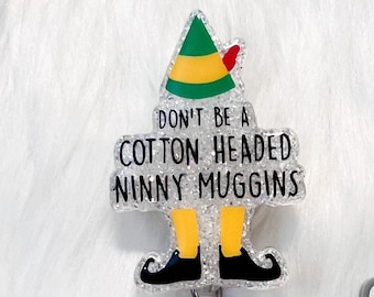 Puede incluir: Un carrete de insignia brillante con un diseño de gorro de elfo verde, amarillo y rojo. El texto en la insignia dice "DON'T BE A COTTON HEADED NINNY MUGGINS". También es visible un logotipo gris de "Rylee Designs".