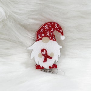 May include: A gnome-shaped badge reel with a red and white color scheme. The gnome's hat is red with white ribbon patterns, and the beard is white with glitter. A red ribbon is on the gnome's beard. The badge reel has a retractable cord and a metal clip.