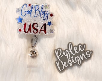 May include: A badge reel with the text "God Bless USA" in red and blue, with star accents. The badge reel is white with glitter and has a clear plastic strap and metal clip. A gray "Rylee Designs" logo is also present.