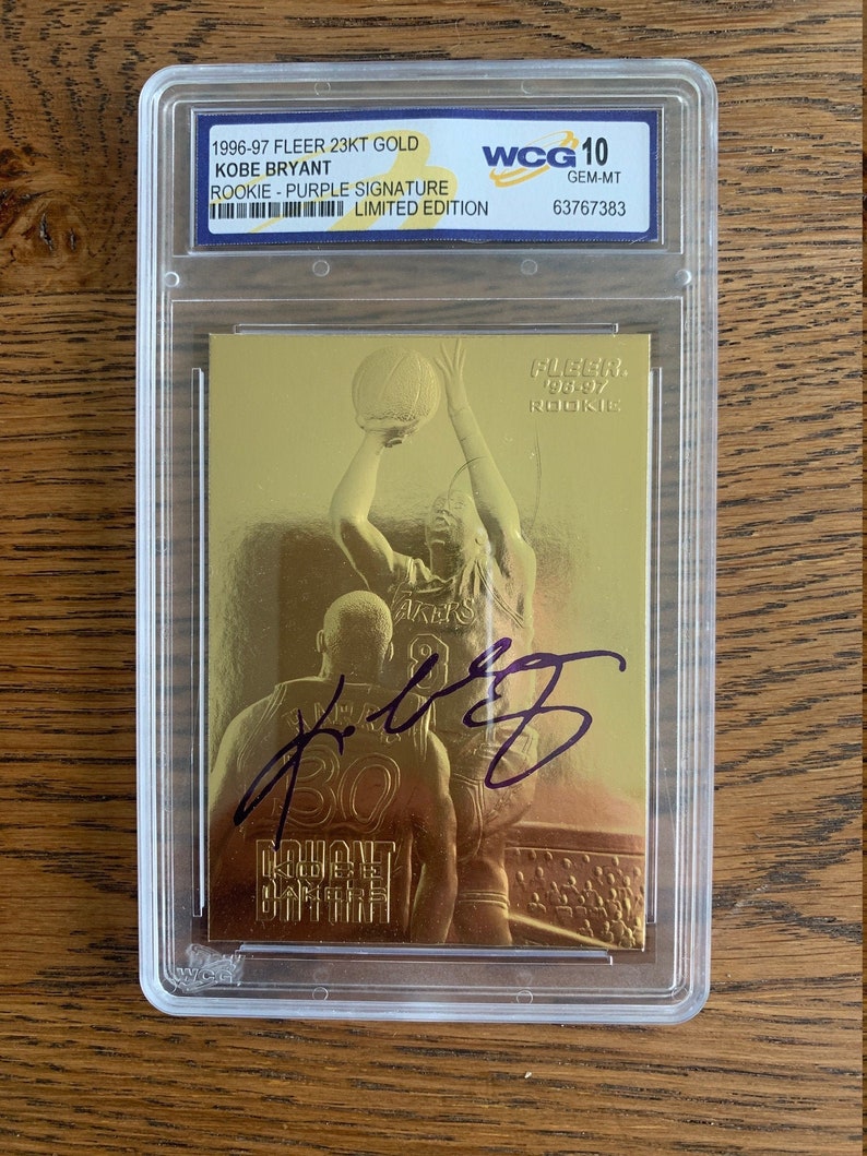 May include: A gold-colored Kobe Bryant rookie trading card with a purple signature. The card is graded GEM-MT 10 by WCG. The card is from the 1996-97 Fleer 23KT Gold series.