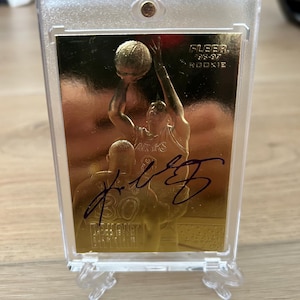 Könnte beinhalten: Eine goldfarbene Basketball-Sammelkarte mit einem Basketballspieler in Aktion. Die Karte ist mit lilafarbener Tinte vom Spieler "Kobe Bryant" signiert. Die Karte ist in einem durchsichtigen Plastik-Schutzgehäuse eingeschlossen und auf einem durchsichtigen Plastikständer ausgestellt. Die Karte stammt aus der Fleer Rookie-Serie von 1996-97.