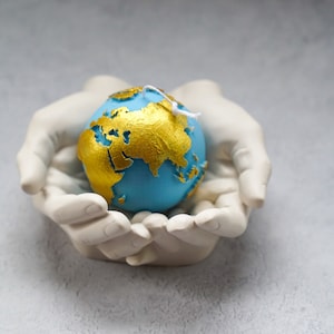 May include: A pair of white ceramic hands holding a blue and gold globe candle. The globe has a textured surface and is decorated with gold leaf.