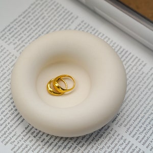Peut inclure: Deux anneaux en or martelé sont présentés dans un porte-bagues en céramique blanche. Le porte-bagues a la forme d'un beignet avec une petite indentation au centre.
