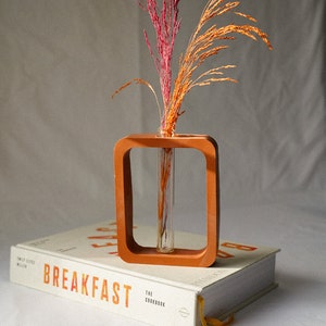 May include: A terracotta vase with a square frame and a glass tube for holding dried flowers. The vase is sitting on top of a white book with the word "BREAKFAST" printed on the cover.