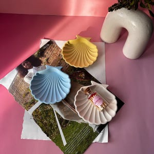 Peut inclure: Trois plats en céramique en forme de coquille de mer en bleu, jaune et blanc. Le plat blanc contient un petit bocal en verre avec des allumettes étiquetées "goblin".