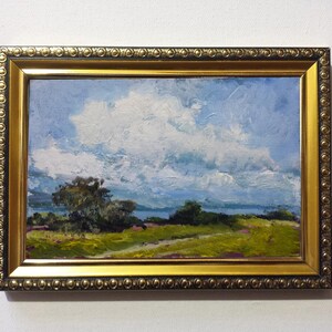 May include: Oil painting of a landscape with a blue sky and large white clouds. The painting features a path through a green field with trees and a body of water in the distance. The painting is in a gold ornate frame.