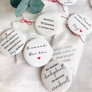 Può includere: Oggetti decorativi circolari bianchi con varie citazioni di testo nero e piccoli accenti a forma di cuore rosso. Alcuni sono legati con uno spago rosso. Un rametto di foglie verdi è sullo sfondo.