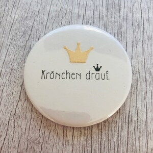 May include: A white, round button with a gold crown graphic and the phrase "Kr&ouml;nchen drauf" in black lettering. A small black crown is also present. The button is on a light gray wooden surface.