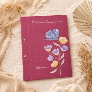 Könnte beinhalten: Eine dunkelrosa Mappe mit zwei silbernen Metallschrauben an der linken Seite. Das Cover zeigt eine florale Illustration mit blauen und gelben Blumen sowie den weißen Schriftzug "Meine Zeugnisse" oben und den Namen "Aenna" in Weiß.