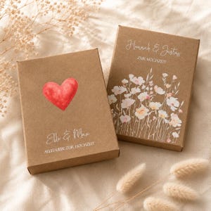 Pode incluir: Duas caixas de presente retangulares de papelão marrom. Uma caixa apresenta um coração vermelho em aquarela e o texto Elli & Max ALLES LIEBE ZUR HOCHZEIT. A outra caixa exibe uma ilustração floral e o texto Hannah & Justus ZUR HOCHZEIT.