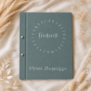 May include: A sage green rectangular scrapbook with a fabric-like texture, featuring two silver metal binding screws on the left side. The cover displays a circular alphabet design surrounding the name Frederik and the text Meine Zeugnisse.