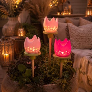 Peut inclure: Trois tuteurs de jardin en forme de tulipe avec des éléments lumineux. Chaque tuteur possède un sommet en céramique rose clair ou rose vif avec des découpes circulaires, monté sur un fin poteau en bois, placé dans une grande jardinière en pierre.