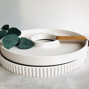 Op de afbeelding: Een wit keramisch serveerblad met een verhoogd midden en een decoratieve, geribbelde basis. De lade heeft een bruin label met de tekst "not for dish". Groene bladeren zijn op de lade geplaatst.