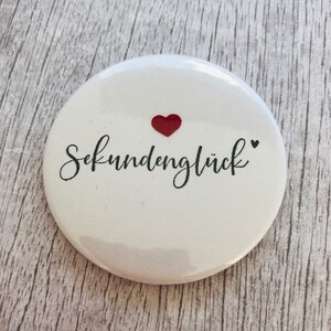 May include: A white, round button with the German word "Sekundengl&uuml;ck" in black script, a small red heart, and a black heart. The button is a simple, yet elegant design.