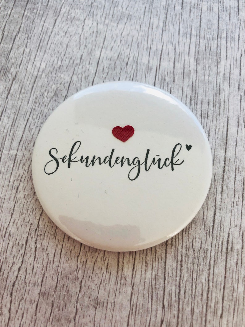 May include: A white, round button with the German word "Sekundengl&uuml;ck" in black script, a small red heart, and a black heart. The button is a simple, yet elegant design.
