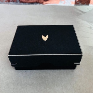 Pode incluir: Uma caixa de presente retangular preta com um adorno dourado em forma de cora&ccedil;&atilde;o na tampa. A caixa tem uma borda prateada e est&aacute; fechada. A caixa &eacute; provavelmente para joias ou pequenos presentes.