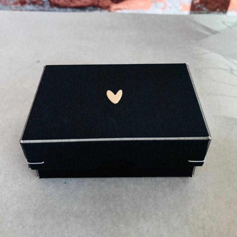 Pode incluir: Uma caixa de presente retangular preta com um adorno dourado em forma de cora&ccedil;&atilde;o na tampa. A caixa tem uma borda prateada e est&aacute; fechada. A caixa &eacute; provavelmente para joias ou pequenos presentes.