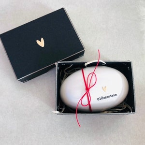 Pode incluir: Um Gl&uuml;cksstein branco, de forma oval, &eacute; apresentado numa caixa preta aberta com um cora&ccedil;&atilde;o dourado. A pedra &eacute; amarrada com uma corda vermelha e apresenta a palavra "Gl&uuml;cksstein". A caixa tem um cora&ccedil;&atilde;o dourado na tampa.