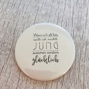 May include: A white, round button with black text. The text reads, "Wenn ich alt bin, will ich nicht jung aussehen, sondern gl&uuml;cklich." The button is on a light wood-grain surface.