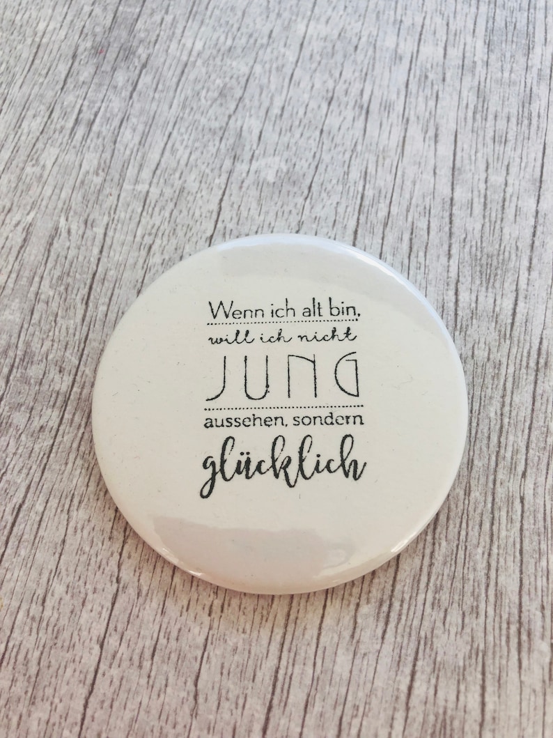 May include: A white, round button with black text. The text reads, "Wenn ich alt bin, will ich nicht jung aussehen, sondern gl&uuml;cklich." The button is on a light wood-grain surface.