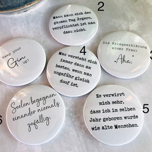 May include: A collection of white round buttons with black text. One button says "Keep your Gin up!" Another says "Aha." Other buttons have German phrases. The buttons are on a light-colored surface.