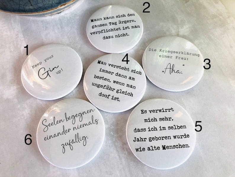 May include: A collection of white round buttons with black text. One button says "Keep your Gin up!" Another says "Aha." Other buttons have German phrases. The buttons are on a light-colored surface.
