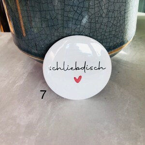 May include: White, round button with the word "schliebdish" in black script and a small red heart. The number 7 is printed in black on the lower left. The button is in front of a gray ceramic pot.