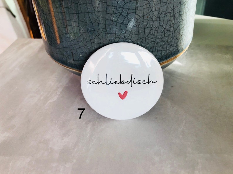 May include: White, round button with the word "schliebdish" in black script and a small red heart. The number 7 is printed in black on the lower left. The button is in front of a gray ceramic pot.
