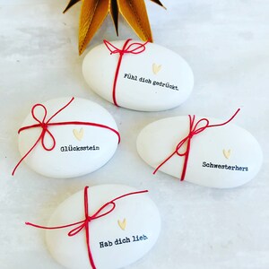Pode incluir: Quatro pedras brancas ovais com barbante vermelho amarrado. Cada pedra tem uma frase alem&atilde; diferente impressa em preto, junto com um pequeno cora&ccedil;&atilde;o dourado. Uma decora&ccedil;&atilde;o dourada em forma de estrela est&aacute; no fundo.