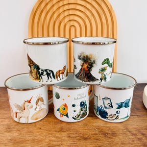 Könnte beinhalten: Fünf weiße Emaille-Tassen mit silbernen Rändern, jede mit einer anderen Illustration. Designs umfassen Pferde, einen Vulkan mit Dinosauriern, ein Einhorn und eine Prinzessin, eine Rakete und einen Astronauten sowie Sportgeräte.