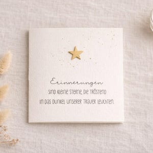 May include: A square, off-white card with a gold star and text in German. The card is decorated with small gold flecks. A lit candle in a glass holder is in the background, along with dried flowers.