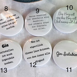 May include: A collection of white, round buttons with black text. Each button features a different phrase or saying. The buttons are arranged on a light-colored surface. The text includes phrases like "Gin Salabim" and "Das Herz will was das Herz will."