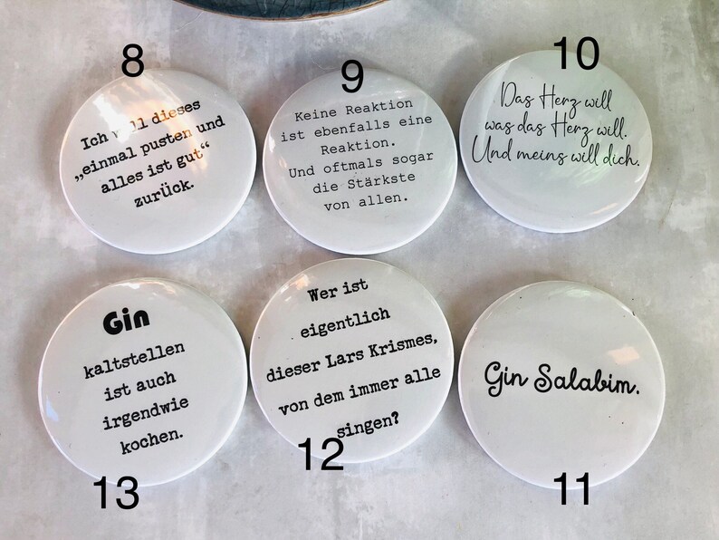 May include: A collection of white, round buttons with black text. Each button features a different phrase or saying. The buttons are arranged on a light-colored surface. The text includes phrases like "Gin Salabim" and "Das Herz will was das Herz will."
