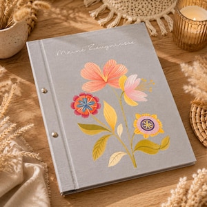 Könnte beinhalten: Ein grauer, stoffbezogener Ordner mit dem in Weiß aufgedruckten Text Meine Zeugnisse. Der Einband zeigt eine bunte Blumenillustration mit rosa, orangefarbenen, gelben und blauen Blütenblättern sowie grünen Blättern. Der Ordner ist mit zwei silbernen Metallnieten befestigt.