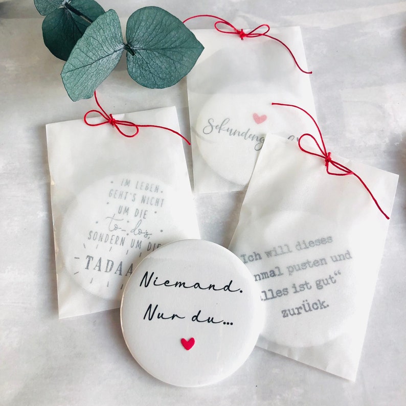 May include: White round objects with text and red heart accents are displayed. One reads "Niemand. Nur du..." with a heart. Others are in bags with text like "Sekundeng" and "Ich will dieses..." tied with red string.