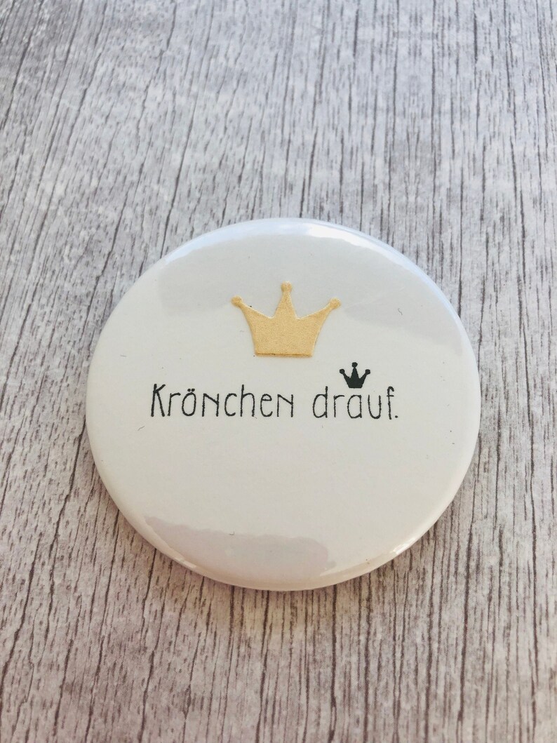 May include: A white, round button with a gold crown graphic and the phrase "Kr&ouml;nchen drauf" in black lettering. A small black crown is also present. The button is on a light gray wooden surface.