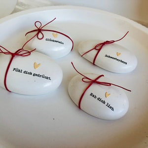 Pode incluir: Quatro pedras brancas ovais com texto em alem&atilde;o e detalhes de cora&ccedil;&atilde;o dourado, amarradas com barbante vermelho. As pedras est&atilde;o inscritas com frases em alem&atilde;o. Est&atilde;o dispostas em um prato branco raso.