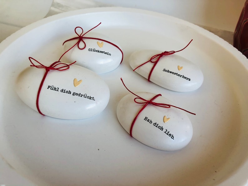 Pode incluir: Quatro pedras brancas ovais com texto em alem&atilde;o e detalhes de cora&ccedil;&atilde;o dourado, amarradas com barbante vermelho. As pedras est&atilde;o inscritas com frases em alem&atilde;o. Est&atilde;o dispostas em um prato branco raso.